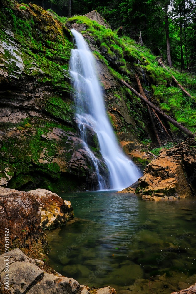 Waterfall cascading over jagged rocks, surrounded by lush greenery in a serene forest landscape.