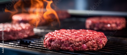 Three beef hamburger patties sizzle on a grill, surrounded by flames. The smell of cooking red meat fills the air, creating a mouthwatering aroma
