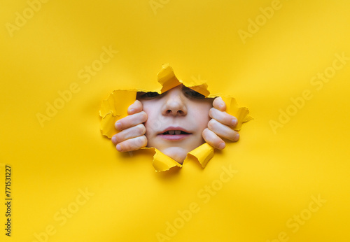 A little girl looks into a torn hole in yellow paper. Hands, nose and mouth. The concept of espionage, children's curiosity, peeping, indifference to what they see. Copy space. Snoopy, inquisitive.