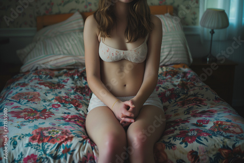 anonymous young woman sitting on bed wearing white underwear, hands clasped concpt of mental health, pregnancy, body issues photo