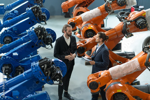 Two professionals in suits engaged in a discussion amid advanced industrial robots in a technology facility.
