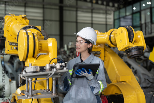 Female industrial engineer in hard hat inspecting new robot arms machinery components while holding a digital tablet in a manufacturing plant.
