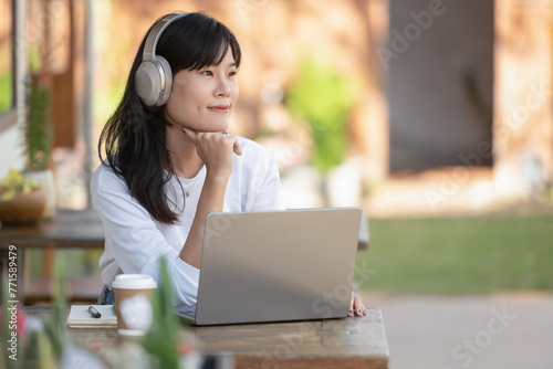 Smiling young adult female with a coffee and headphones at an outdoor cafe, using a laptop.