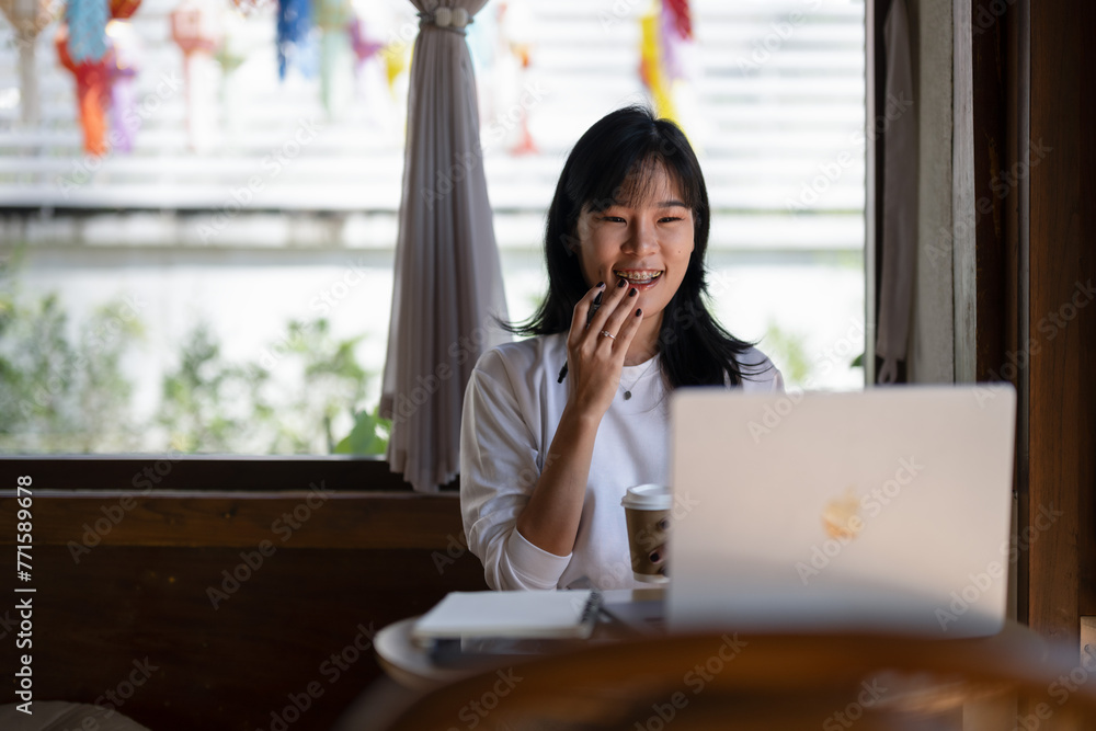 Fototapeta premium .Young Asian woman enjoying coffee while having a pleasant video call on a laptop in a coffee shop.