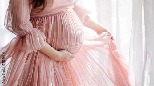 A pregnant woman wearing a pink dress stands in front of a window, looking outside