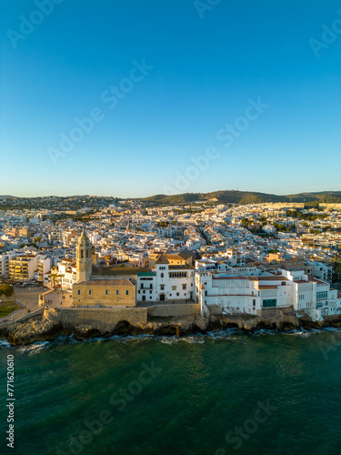 Drone point of view of Sitges City at sunset. Panoramic view of all city. Waves splashing the rocks and buildings. Travel destination. Beautiful warm colours at sunset, reflecting on buildings. 