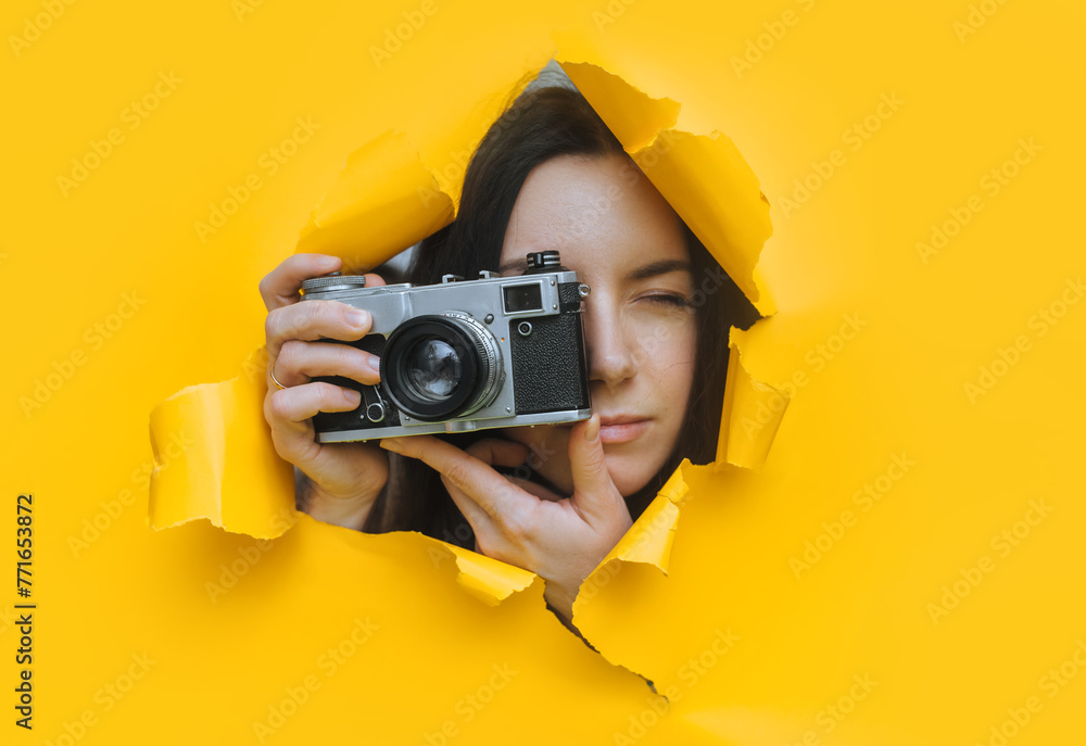 A young paparazzi girl with a rare SLR camera looks out from her hiding place and carefully watches what is happening. Yellow paper hole. Tabloid press. Looking for a subject for stock photos. - obrazy, fototapety, plakaty 