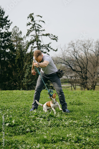 Caucasian man plays toy rope tug-of-war with a dog on green grass. An active spring walk in park of male owner and jack Russell terrier puppy. The concept of a healthy lifestyle and pet care.