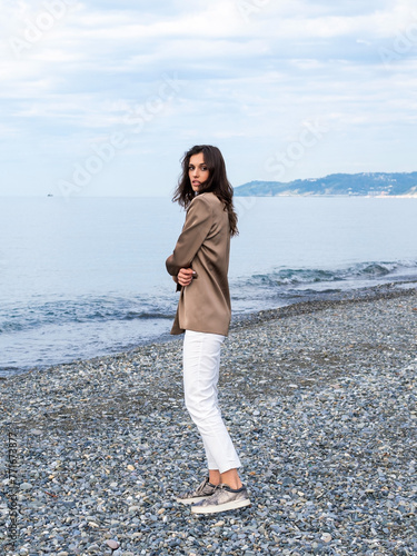 beautiful brunette in a brown satin jacket, white trousers and sneakers walks along the beach in winter. Attractive slim woman looking over her shoulder