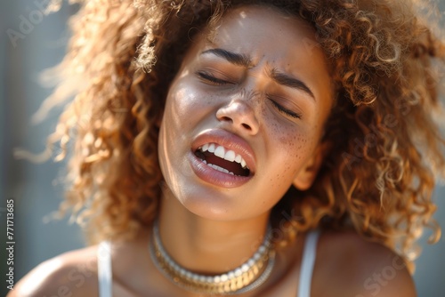 A curly-haired young woman with eyes closed and a pained expression, possibly from bright light or discomfort