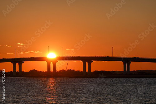 Setting sun blocked by a long bridge with an orange sky and reflection on the smooth bay water