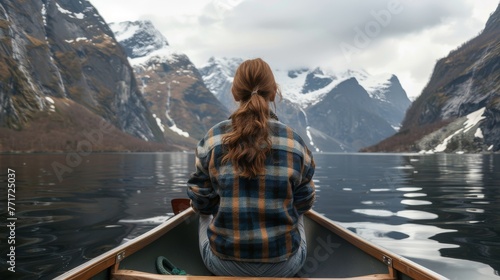  A woman sits in a boat, facing a mountain range with snow-capped peaks in the backdrop of a tranquil lake