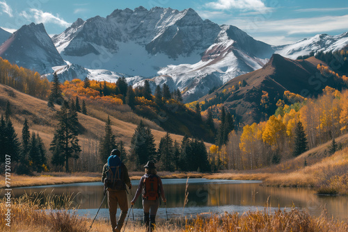 A couple hiking in the mountains surrounded by autumn foliage and snowcapped peaks. They carry backpacks and walking sticks as they walk along an alpine lake. We see their connection with nature.