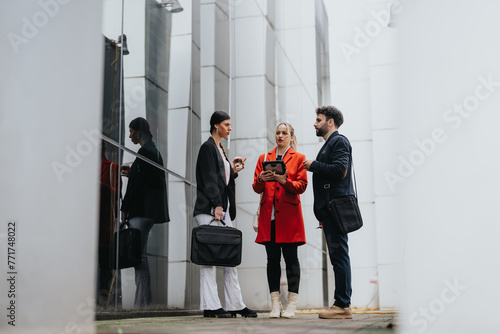 Three colleagues in smart attire engage in a discussion with digital tablet and coffee outside an office building.