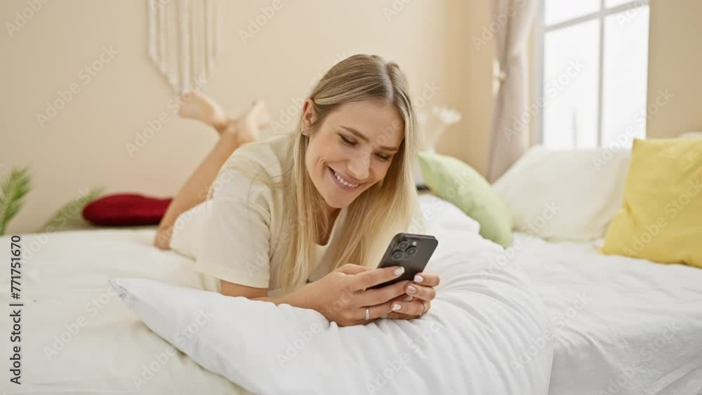 Confident young blonde woman enjoying morning in bed, smilingly using smartphone in a comfortable bedroom setting
