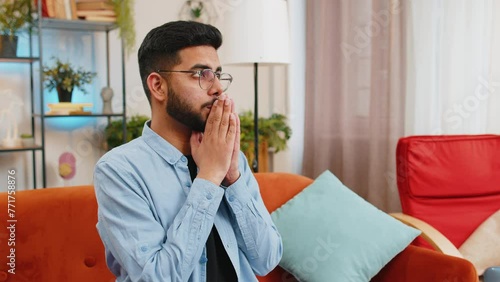 Tensed Hispanic man sits on sofa at home folding palms together pray ask for help thinks feel stressed looks desperate by life problems. Worried upward Indian guy praying making wish begging apology