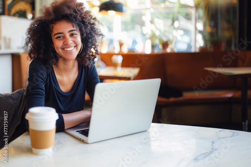 Cheerful woman works on laptop at a table with a coffee cup beside her, in a bright space with natural light, exuding a relaxed vibe. © mimi