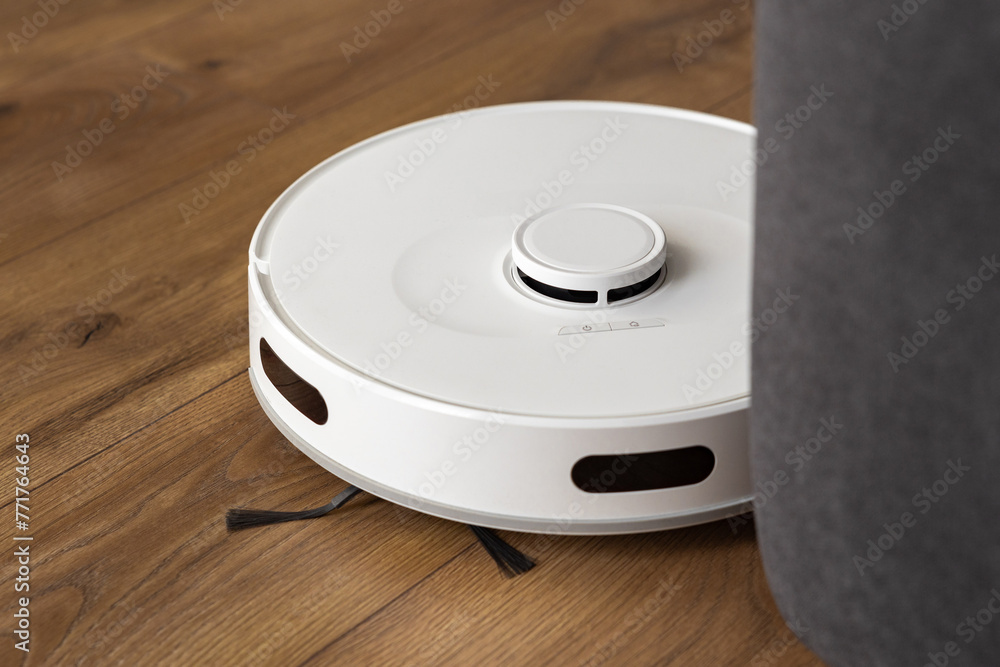 A white robot hoover cleans a dark oak wood laminate on the floor. collects dust near the sofa in difficult places. automatic management of modern technologies. infrared sensors control A white robot hoover cleans a dark oak wood laminate on the floor. collects dust near the sofa in difficult places. automatic management of modern technologies. infrared sensors control