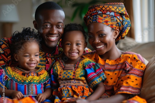 Portrait of happy African family dressed in bright colorful ethnic style clothes posing on a couch at home. Cheerful young parents cuddling their cute kids, smiling broadly and looking at camera.