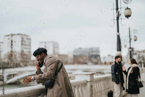 A well-dressed man in a coat and cap engaged in a conversation on his mobile phone while checking his watch on an bridge.