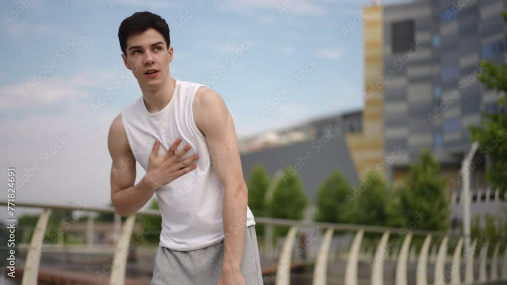 Slow motion. A young guy stopped during his morning jog to catch his breath. The guy rubbed his hands together and wiped sweat from his forehead, while standing outdoors in a city park against the Slow motion. A young guy stopped during his morning jog to catch his breath. The guy rubbed his hands together and wiped sweat from his forehead, while standing outdoors in a city park against the