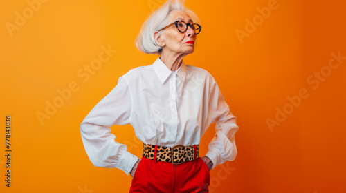 Old woman with white hair with round glasses wearing white blouse, red pants and leopard print belt standing isolated over orange background.