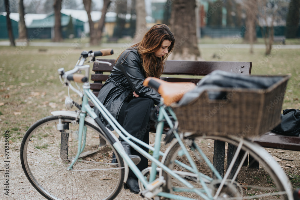 Obraz premium Confident young businesswoman taking a break outdoors, exuding elegance and style while sitting on a park bench beside a bicycle.