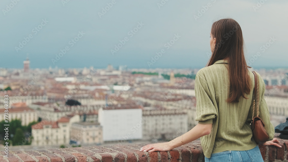 Young woman with brown hair wearing an olive green sweater looks at the city from the observation deck, Back view