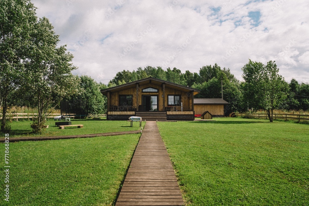 Wooden Cabin with Pathway in Green Landscape. A wooden cabin retreat is nestled in a lush green landscape, complete with a welcoming boardwalk and surrounding trees.