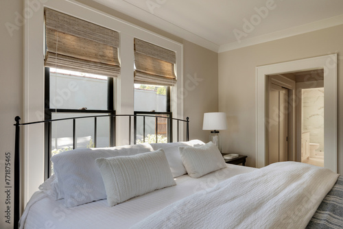 A sun-drenched bedroom in a traditional cottage with a comfortable bed positioned between two windows, allowing ample sunlight to filter in. A stylish headboard frames the bed.