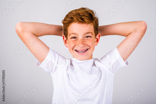 Head and shoulders portrait of a preteen boy with freckles and auburn hair, smiling with braces, while posing with his hands behind his head