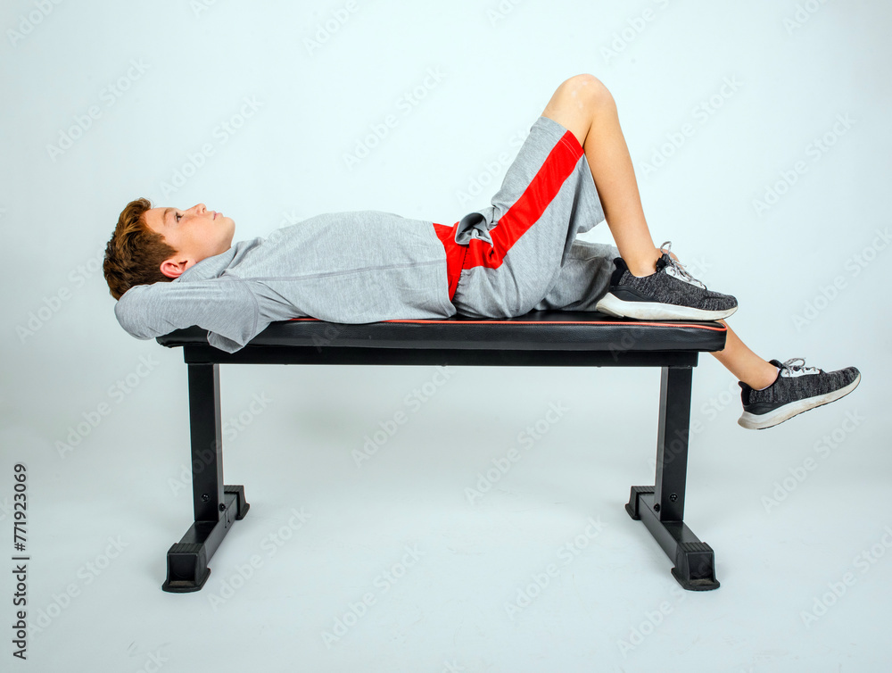 Obraz premium Young preteen male athlete in a grey sweatshirt and shorts, lying back on a bench, hands behind head, looking up and thinking about the game