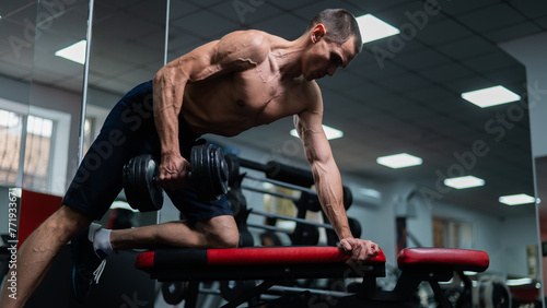 Shirtless man doing dumbbell row to waist on bench at gym. 