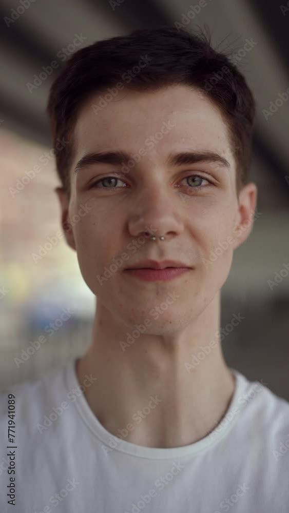 Vertical video. Close-up. A young athletic guy in a white T-shirt looks at the camera and cute smiles, while standing outdoors on a summer day under a city bridge Vertical video. Close-up. A young athletic guy in a white T-shirt looks at the camera and cute smiles, while standing outdoors on a summer day under a city bridge