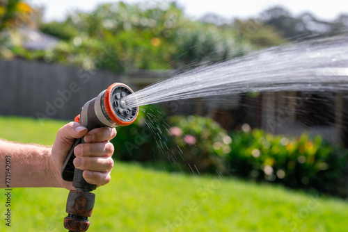 Garden hose with nozzle. Man's hand holding spray gun and watering plants, spraying water on grass in backyard.