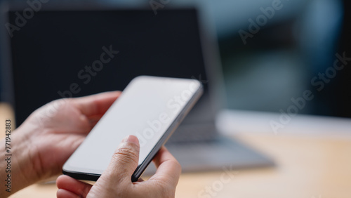 A person is holding a cell phone in their hand, with a laptop in the background. Concept of productivity and focus, as the person is likely working on their phone or laptop