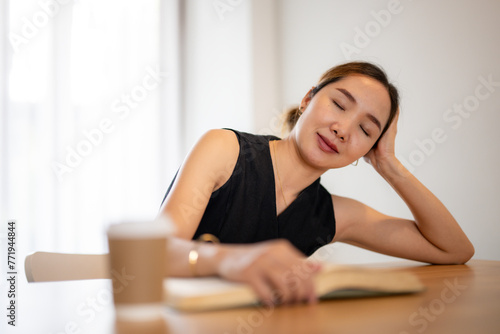 Friendly Asian woman smiles at the camera, resting her arms on a wooden desk with an open book and glasses, portraying a comfortable study environment...