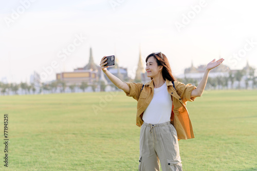 Traveler asian woman in her 30s making a livestream or selfie on smartphone while explores Wat Phra Kaew emerald Buddha. Share the wonders of Thai heritage through her journey.