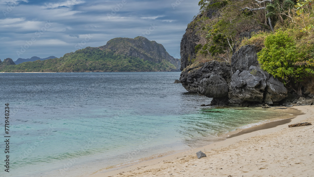 A secluded sandy beach on a tropical island. A calm turquoise ocean. Green vegetation on picturesque coastal cliffs. A mountain on a background of blue sky and clouds. Philippines. Palawan. El Nido