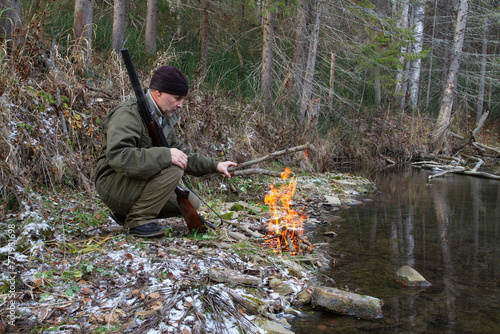 A hunter puts firewood on a campfire on the bank of a forest river