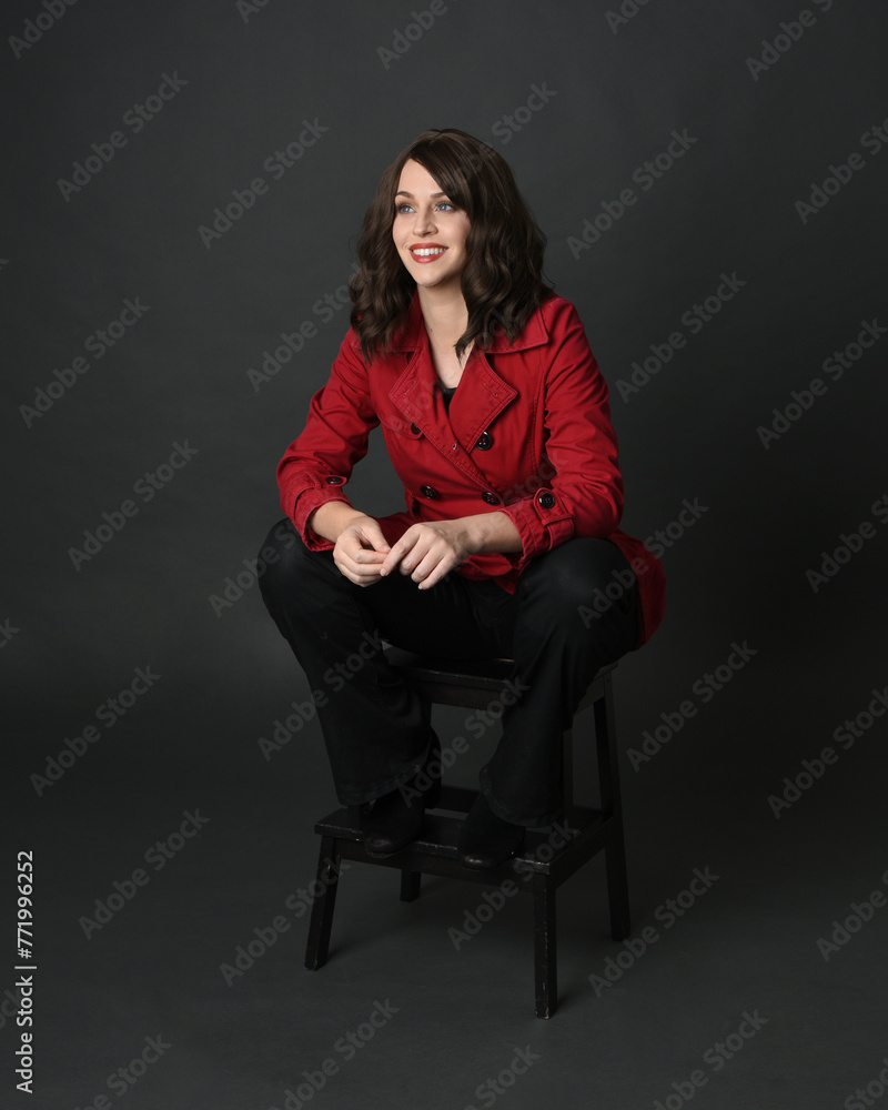 full length portrait of beautiful brunette woman model, wearing red trench coat jacket with black leather pants. sitting pose with hand gesture on dark grey studio background. full length portrait of beautiful brunette woman model, wearing red trench coat jacket with black leather pants. sitting pose with hand gesture on dark grey studio background.