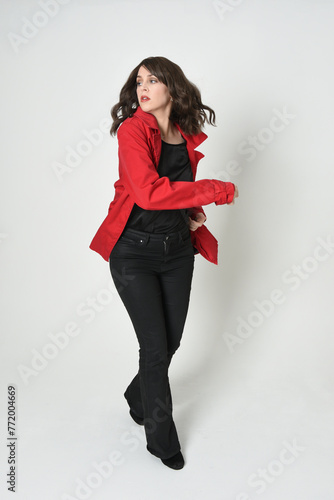 full length portrait of beautiful brunette woman model, wearing red trench coat jacket leather pants. standing pose, walking towards the camera. isolated on white studio background.