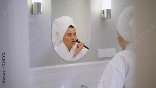 A girl is putting on makeup in front of the mirror. A young girl does her makeup. White bathroom. A girl is applying makeup to her face with a brush. A girl is going to a meeting after a shower. High