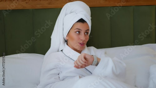 A young girl lies in bed and speaks through the clock. Young girl resting in bed and chatting on the phone. Apple watch. Communication. Weekend day. White bed. A young brunette girl Records a voice