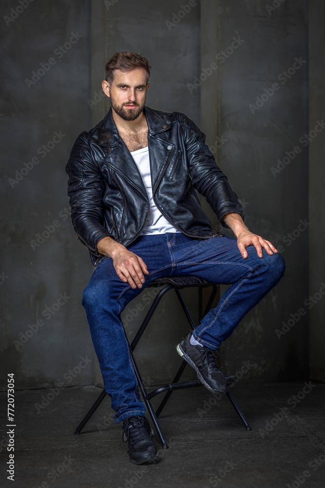 Modern young man with a stylish hairstyle, wearing a cool black leather jacket and sitting on a chair on a dark background Modern young man with a stylish hairstyle, wearing a cool black leather jacket and sitting on a chair on a dark background