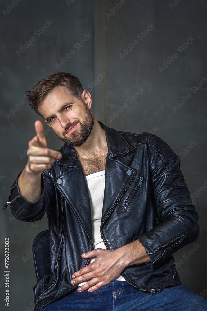Modern young man with a stylish hairstyle, wearing a cool black leather jacket and sitting on a chair on a dark background Modern young man with a stylish hairstyle, wearing a cool black leather jacket and sitting on a chair on a dark background