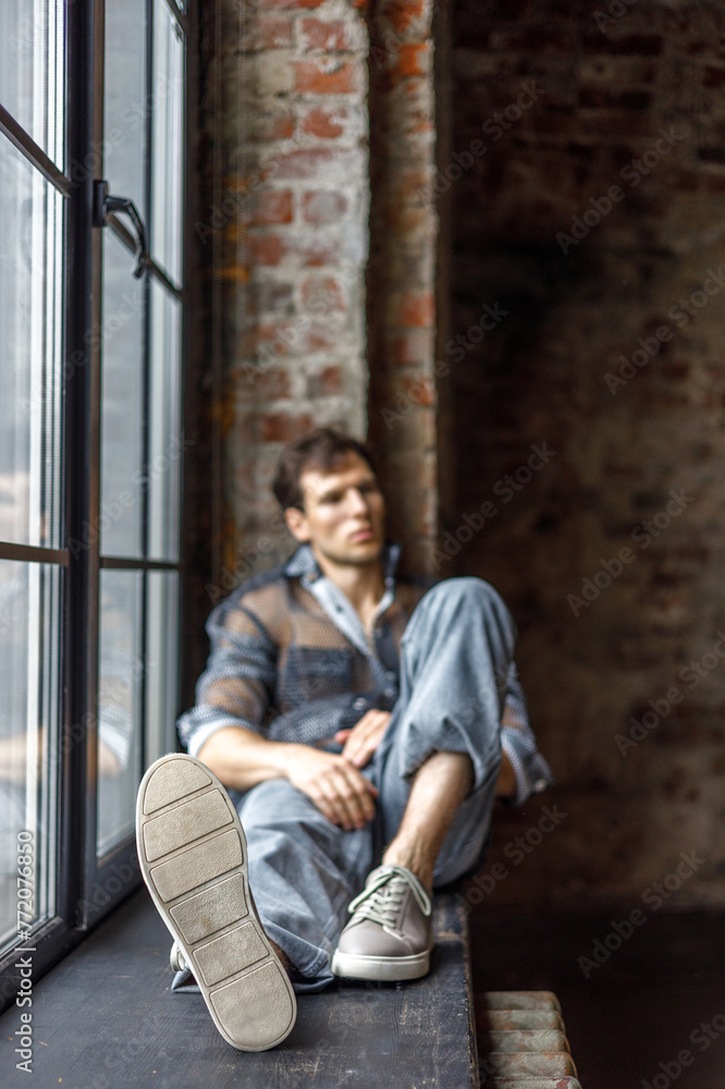 A modern young man with a stylish hairstyle sits on a windowsill near a window on a dark background A modern young man with a stylish hairstyle sits on a windowsill near a window on a dark background