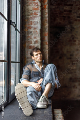 A modern young man with a stylish hairstyle sits on a windowsill near a window on a dark background