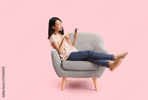 Beautiful Asian woman using mobile phone in grey armchair on pink background