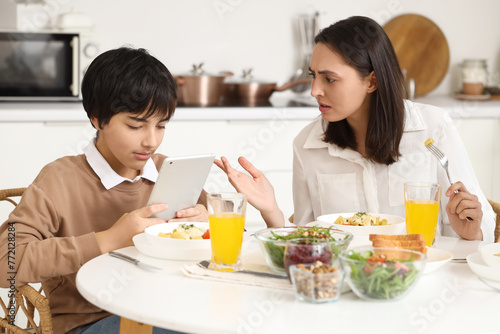 Teenage boy using tablet computer and his angry mother during dinner at table in kitchen. Family problem concept